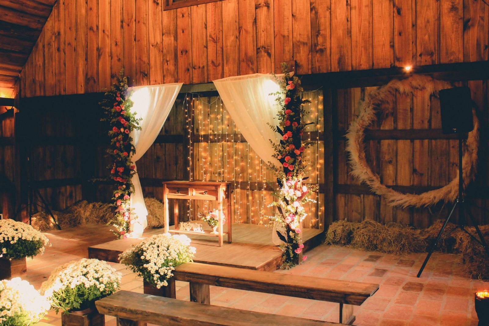 A wedding ceremony set up in a rustic barn with white drapes, flowers, hay bales and a wreath on the wall