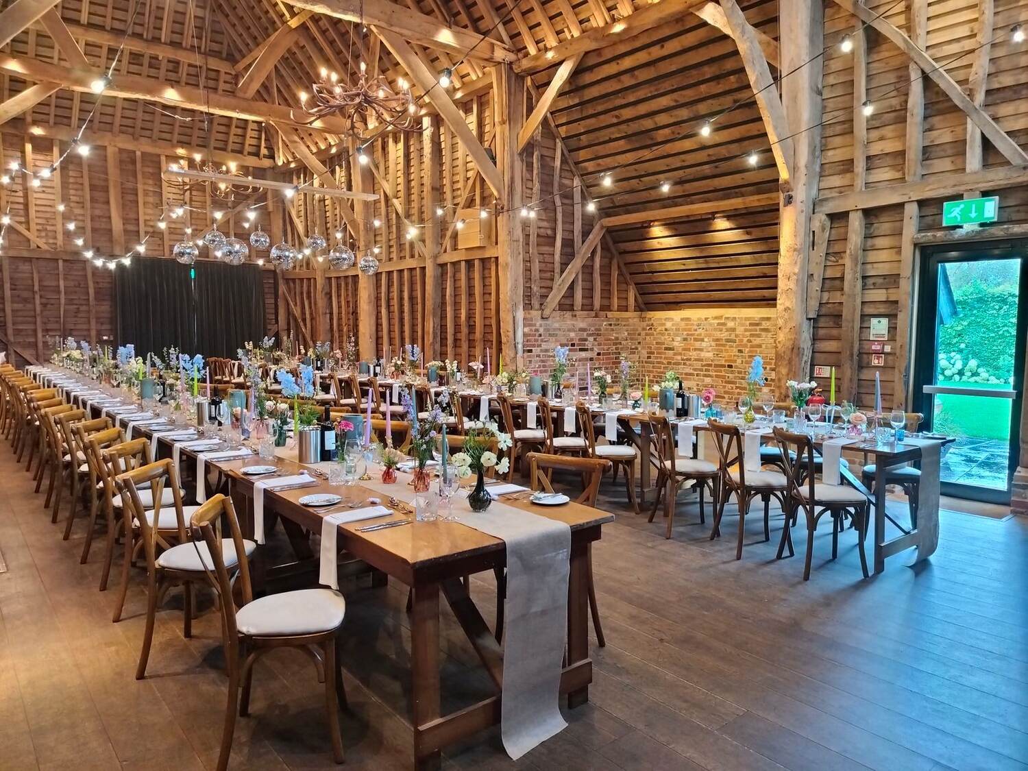 A long banquet table set with flowers in a timber-framed barn strung with lights
