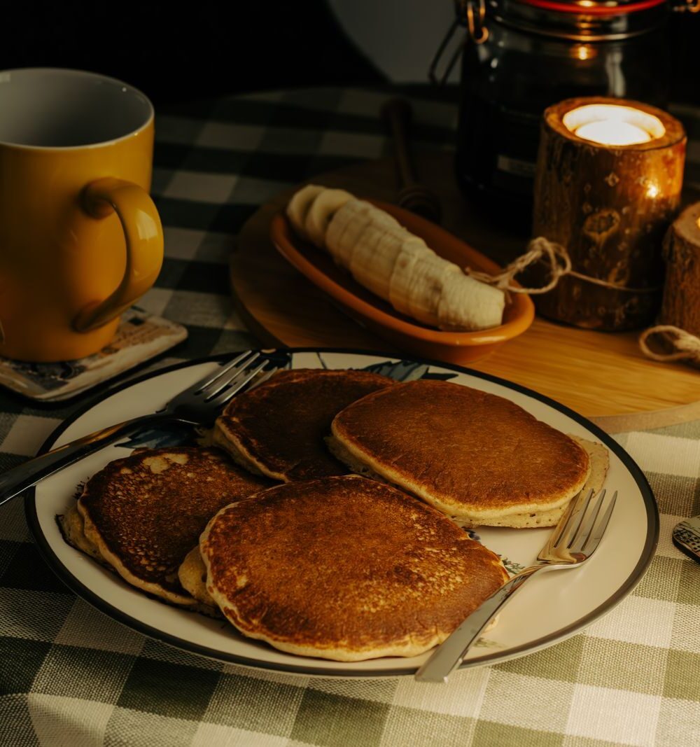 A plate of pancakes on a green checked tablecloth beside two mugs of coffee and candles in wooden holders