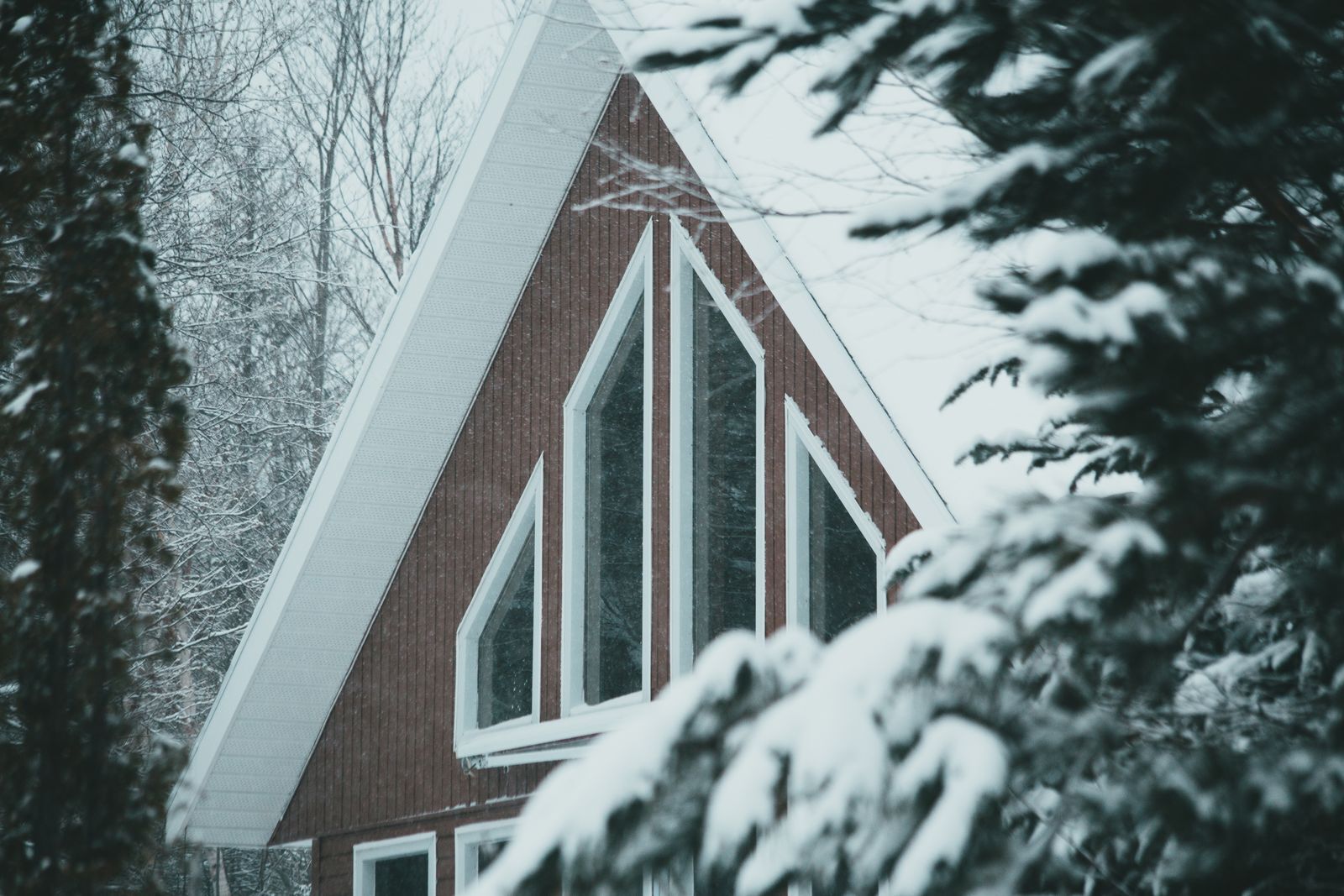 A steep-roofed wooden cottage with tall windows, covered in snow among evergreens