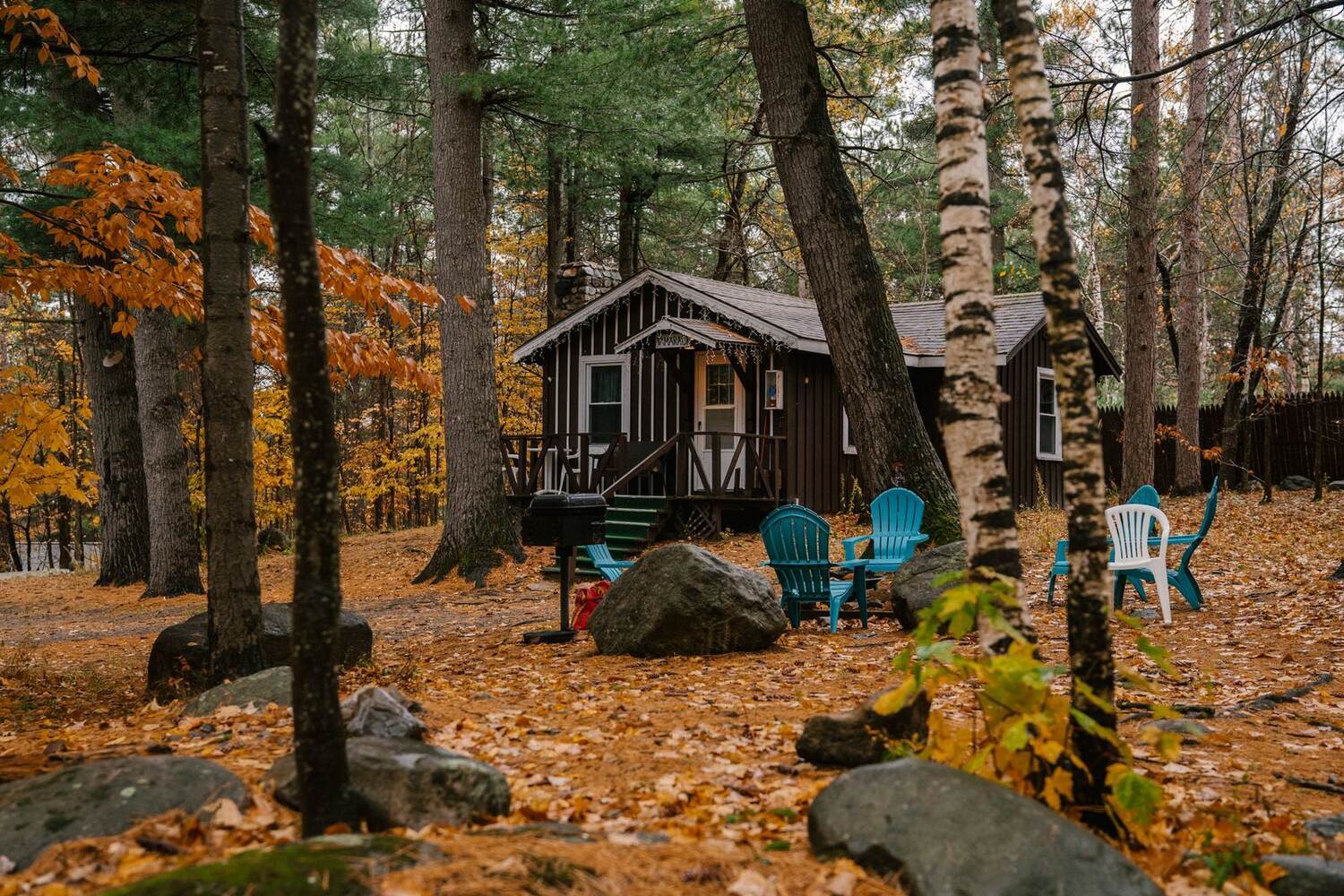 Adirondack chairs in a ring around a fire pit in front of a log cabin among autumn trees