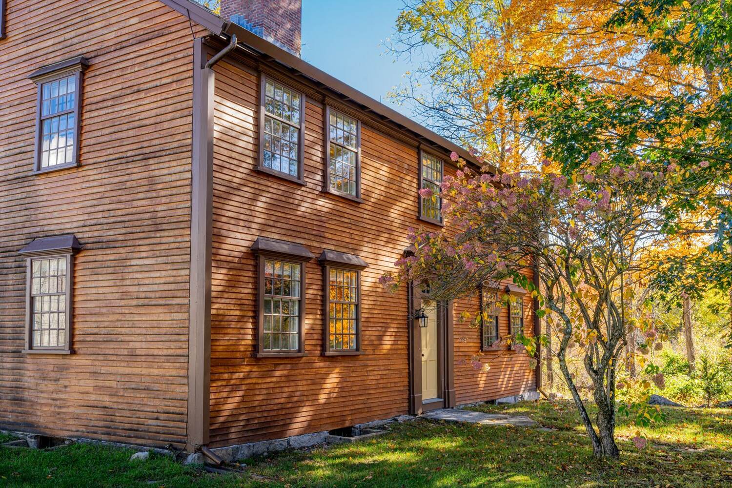 A two-story wooden inn with tall paned windows in warm autumn light, a maple turning gold beside it
