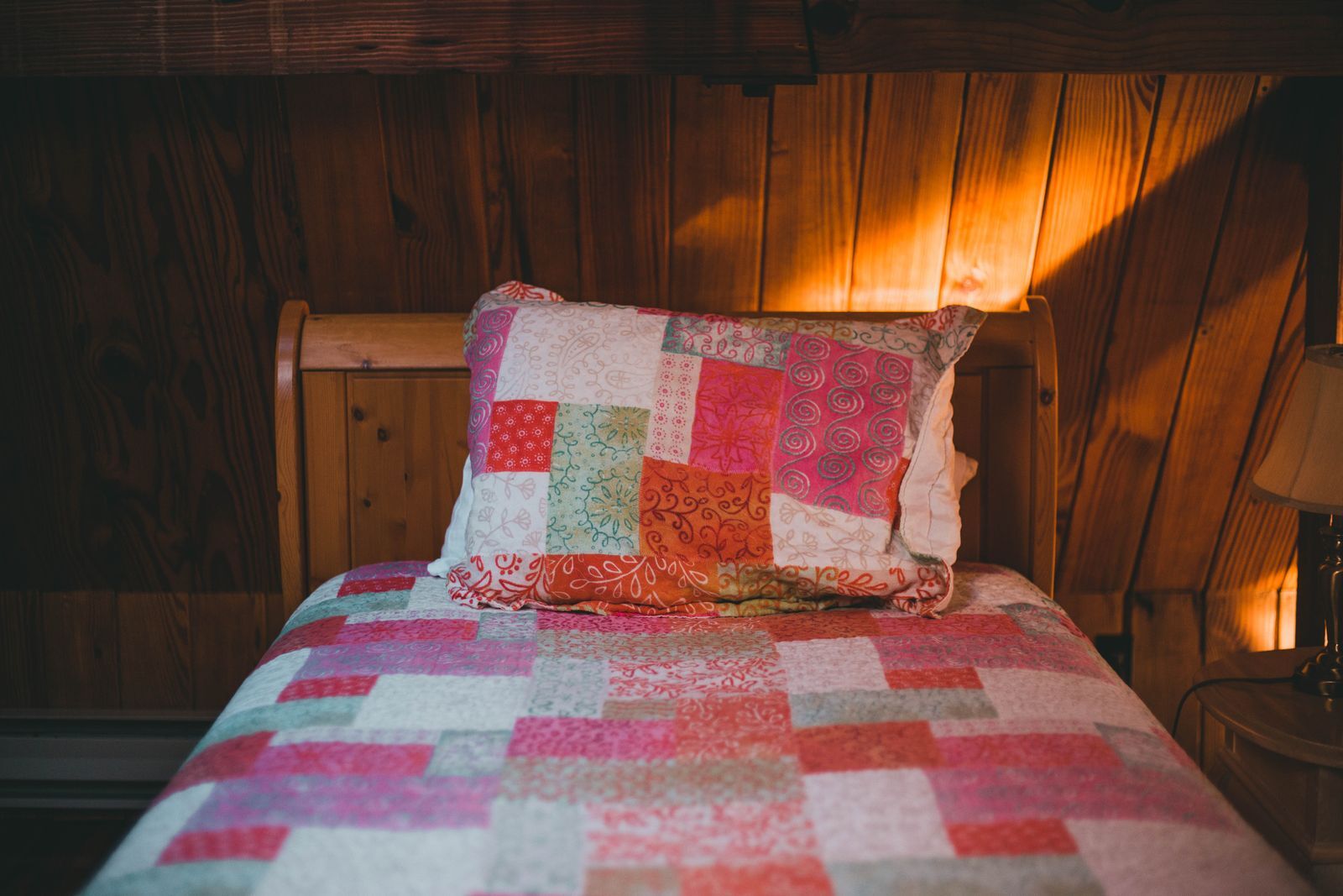 A knotty-pine bedroom with a bed under a red and pink patchwork quilt in afternoon sun