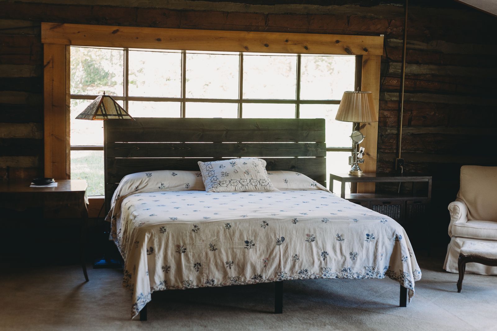 A bedroom with a big window over the bed, wood-paneled walls and a lamp on either side
