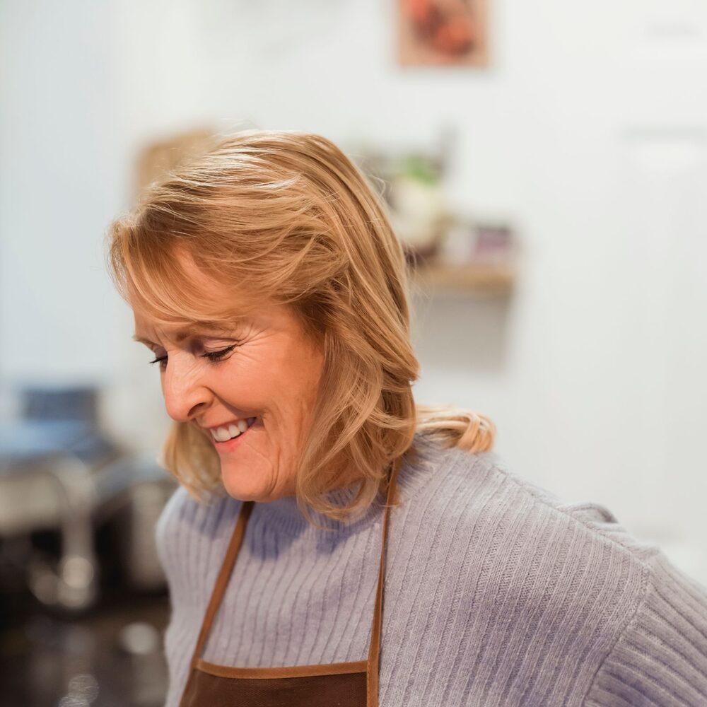 A smiling woman with shoulder-length blond hair in a brown apron, looking down in a bright kitchen