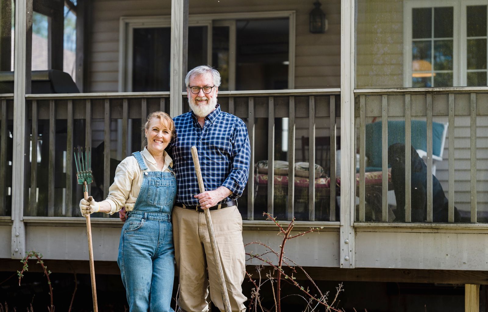A smiling couple in their sixties on the porch of a wooden house, he in a plaid shirt, she holding a garden tool